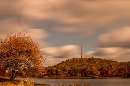A tall monument on a hill surrounded by autumn trees, reflecting in a calm lake under a cloudy sky.