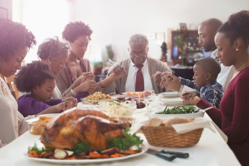 A family gathers around a table, holding hands in prayer before a festive meal, featuring a turkey and various dishes.