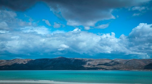 A serene landscape featuring a calm turquoise sea, mountains in the background, and a dramatic sky with clouds.