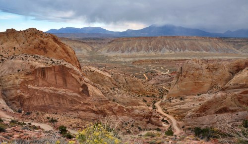 A panoramic view of rugged, colorful rock formations and winding dirt roads under a cloudy sky.