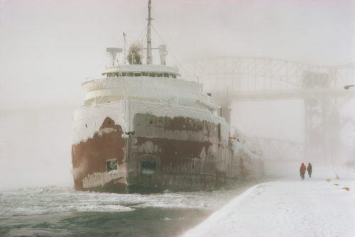 A large ship covered in ice navigates through a snowy landscape, with two people walking along the frozen shore.