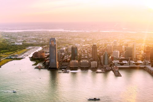 Aerial view of a city skyline at sunset, with water and boats in the foreground and green areas along the shore.