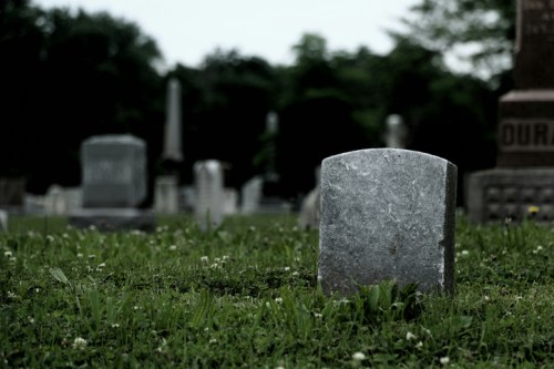 A weathered gravestone stands in a grassy cemetery, surrounded by blurred tombstones and trees in the background.