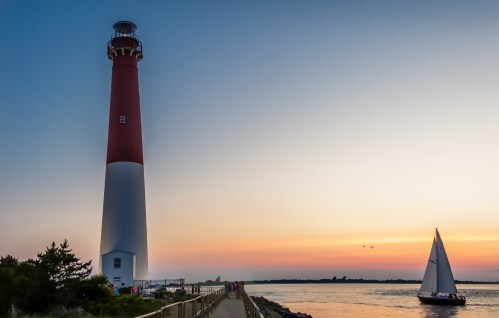 A red and white lighthouse stands by the water at sunset, with a sailboat passing by and a serene sky in the background.