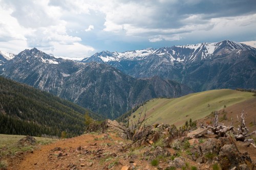A panoramic view of snow-capped mountains under a cloudy sky, with green hills in the foreground.