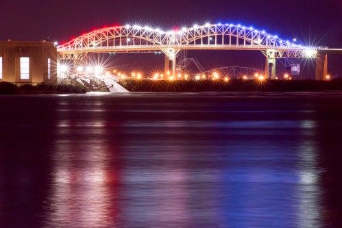 A brightly lit bridge at night, adorned with red, white, and blue lights, reflecting on the calm water below.