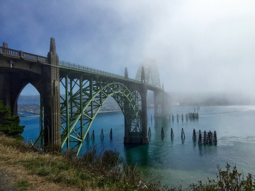 A green bridge arches over calm waters, partially shrouded in fog, with remnants of old piers visible nearby.
