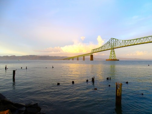 A long bridge stretches over calm water, with clouds and mountains in the background, reflecting golden light.