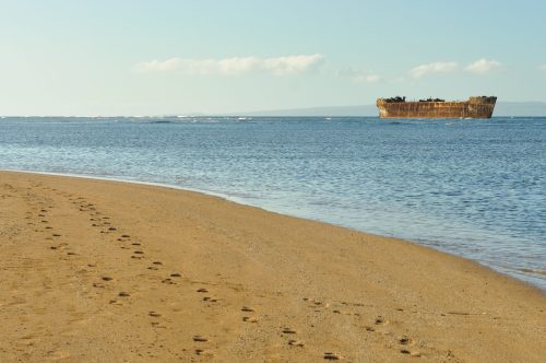 A sandy beach with footprints leading to calm waters, featuring a rusted shipwreck in the distance under a clear sky.