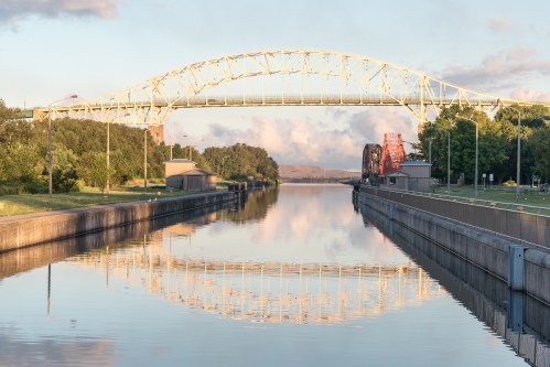 A bridge spans a calm canal, reflecting in the water, surrounded by trees and buildings under a clear sky.