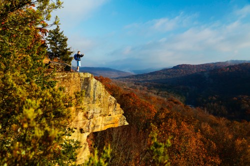A person stands on a cliff edge, taking photos of a colorful autumn landscape with rolling hills in the background.