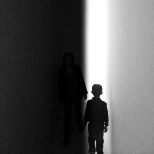 A silhouette of a woman and a child walking through a narrow, dimly lit corridor with a bright light at the end.