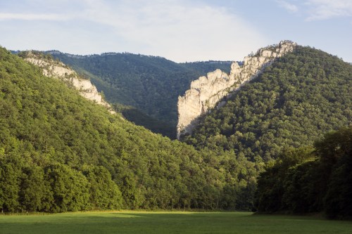 A scenic view of steep, rocky cliffs surrounded by lush green mountains and a grassy valley below.