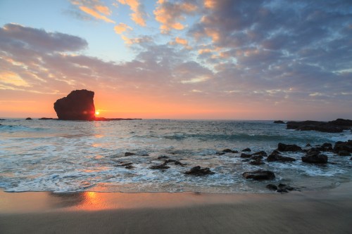 Sunset over the ocean with a rocky outcrop silhouetted against colorful clouds and gentle waves on the shore.