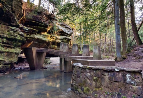 A serene forest scene featuring stone pillars over a calm stream, surrounded by lush greenery and rocky cliffs.
