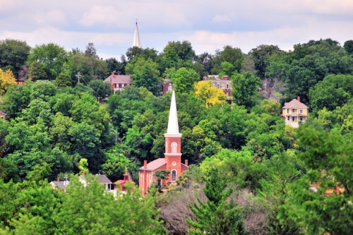 A red brick church with a tall white steeple surrounded by lush green trees and houses on a hillside.