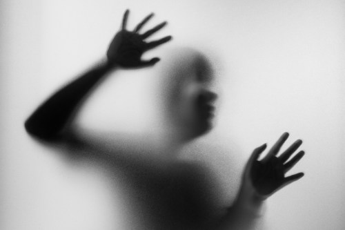 A blurred silhouette of a person pressing against a frosted surface, hands raised in a gesture of struggle or longing.