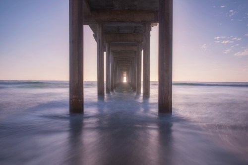 A serene view of a pier extending over calm waters, with soft waves and a pastel sky in the background.