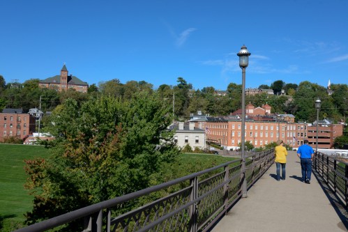 Two people walk along a bridge with a view of buildings and trees under a clear blue sky.