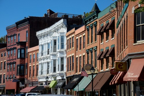 Historic buildings line a street, featuring a mix of brick and white facades, with awnings and an American flag visible.