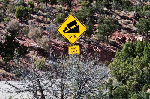 A warning sign indicating a 12% grade slope ahead, with a smaller sign below stating "Next 1 Mile." Surrounding desert landscape.