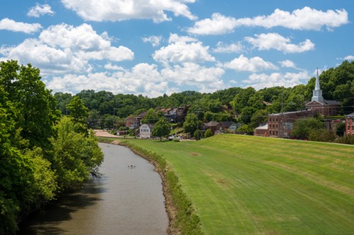 A serene river flows beside lush green fields and quaint buildings under a bright blue sky with fluffy clouds.