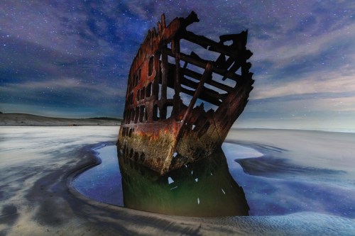 A shipwreck stands partially submerged in water, reflecting the starry night sky and surrounded by sandy terrain.