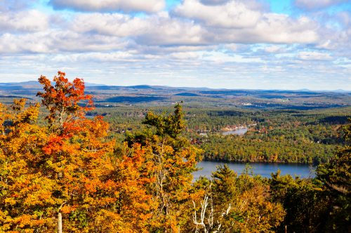 A scenic view of a colorful autumn landscape with trees, a lake, and distant mountains under a partly cloudy sky.
