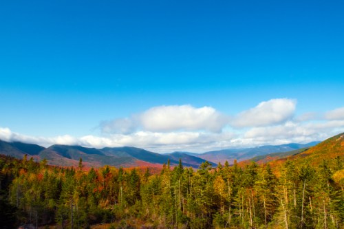 A panoramic view of colorful autumn foliage in the mountains under a clear blue sky with fluffy white clouds.