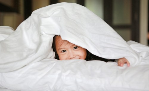 A child peeks out from under a white blanket, smiling playfully in a cozy setting.