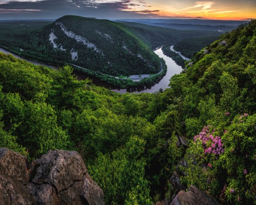 A panoramic view of a lush green valley with a winding river at sunset, surrounded by mountains and blooming flowers.