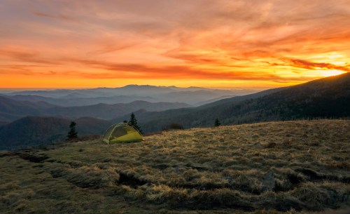 A yellow tent sits on a grassy hillside at sunset, with mountains and a colorful sky in the background.