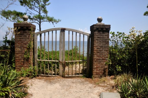 A weathered wooden gate between brick pillars opens to a view of the ocean, surrounded by greenery and sandy path.