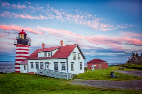 A striped lighthouse beside a white house and red building, set against a colorful sky and ocean backdrop.