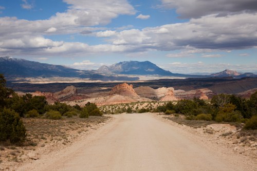 A dirt road leads through a rugged landscape with mountains and clouds in the background.