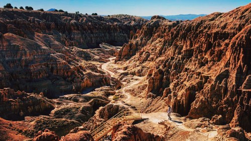 A person walks along a path in a rugged, colorful canyon landscape under a clear blue sky.