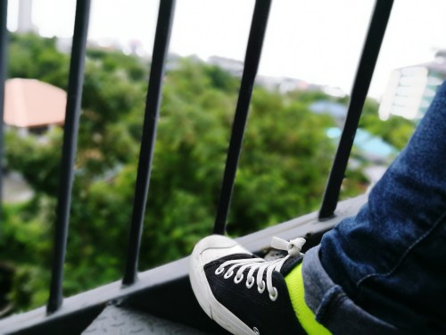 A close-up of a black sneaker with a bright green sock, resting on a balcony railing with greenery in the background.
