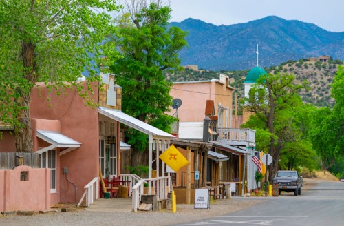 A quaint street in a small town with adobe buildings, trees, and mountains in the background. A vehicle is parked nearby.