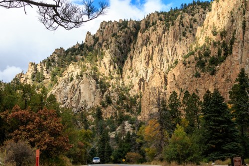 A scenic view of towering rocky cliffs surrounded by trees and autumn foliage, with a car on a winding road below.