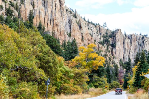 A scenic road lined with colorful autumn trees and towering rock cliffs in the background.