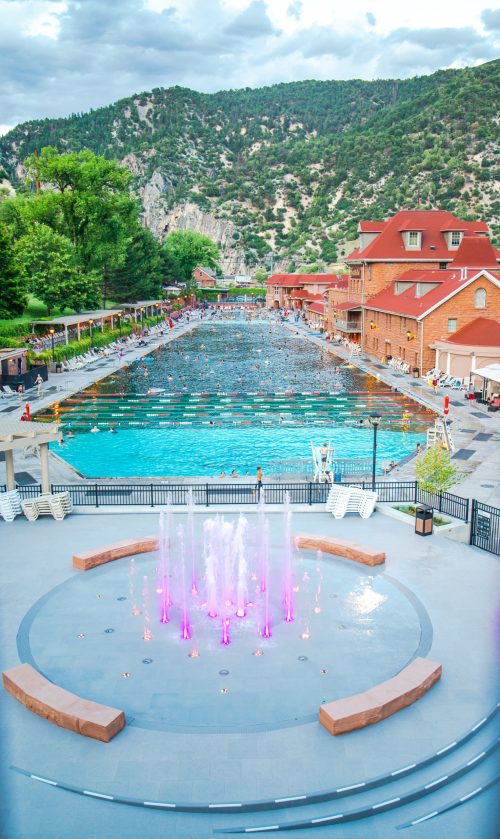 A scenic view of a large swimming pool with colorful fountains, surrounded by mountains and historic buildings.