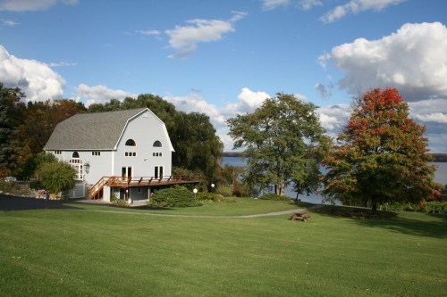 A serene lakeside scene featuring a gray house, lush green lawn, and colorful autumn trees under a partly cloudy sky.