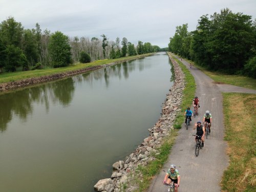Cyclists ride along a canal path lined with trees, reflecting a cloudy sky in the calm water.