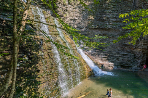 A serene waterfall cascades over rocky cliffs into a clear pool, with people enjoying the natural beauty nearby.