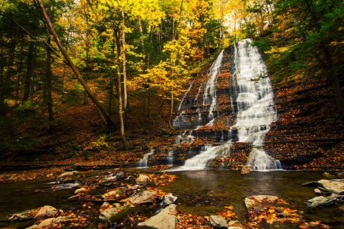 A serene waterfall cascades down rocky steps, surrounded by vibrant autumn foliage and a tranquil stream below.