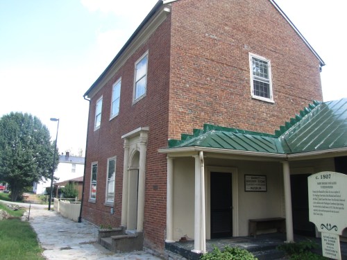 Historic brick building with a green metal roof, featuring large windows and a sign, surrounded by greenery.