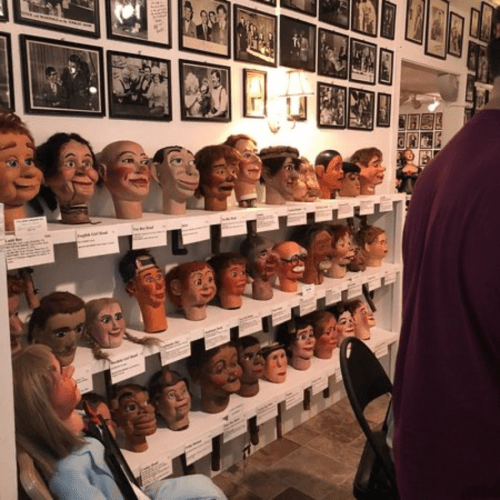 A wall display of various ventriloquist dummies and heads, with black-and-white photos in the background.