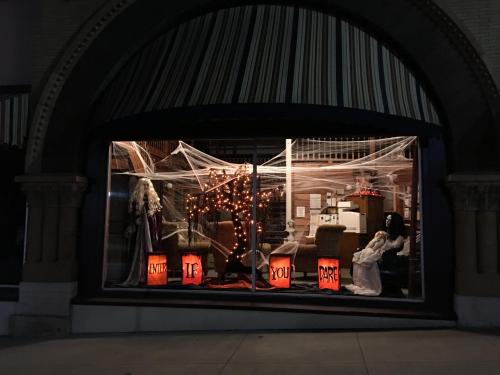 A spooky Halloween display in a window featuring cobwebs, eerie figures, and glowing orange signs.