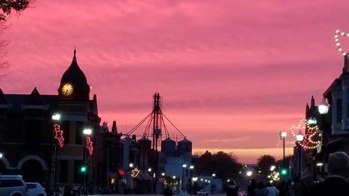 A vibrant sunset casts pink hues over a historic town street, with decorative lights and a clock tower in view.