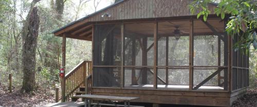 A wooden cabin with a screened porch, surrounded by trees in a natural setting. A picnic table is in front.